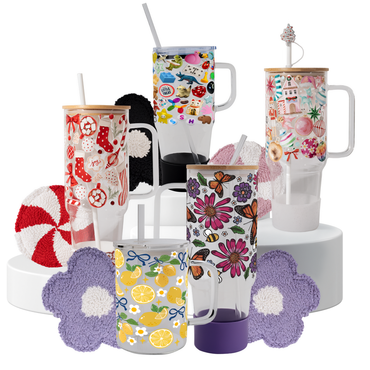 A set of six clear glass tumblers with handles and straws, decorated with various colorful patterns including butterflies, flowers, lemons, candy canes, and retro motifs. The tumblers are arranged on white cylindrical pedestals with matching purple and pink flower-shaped coasters in front.