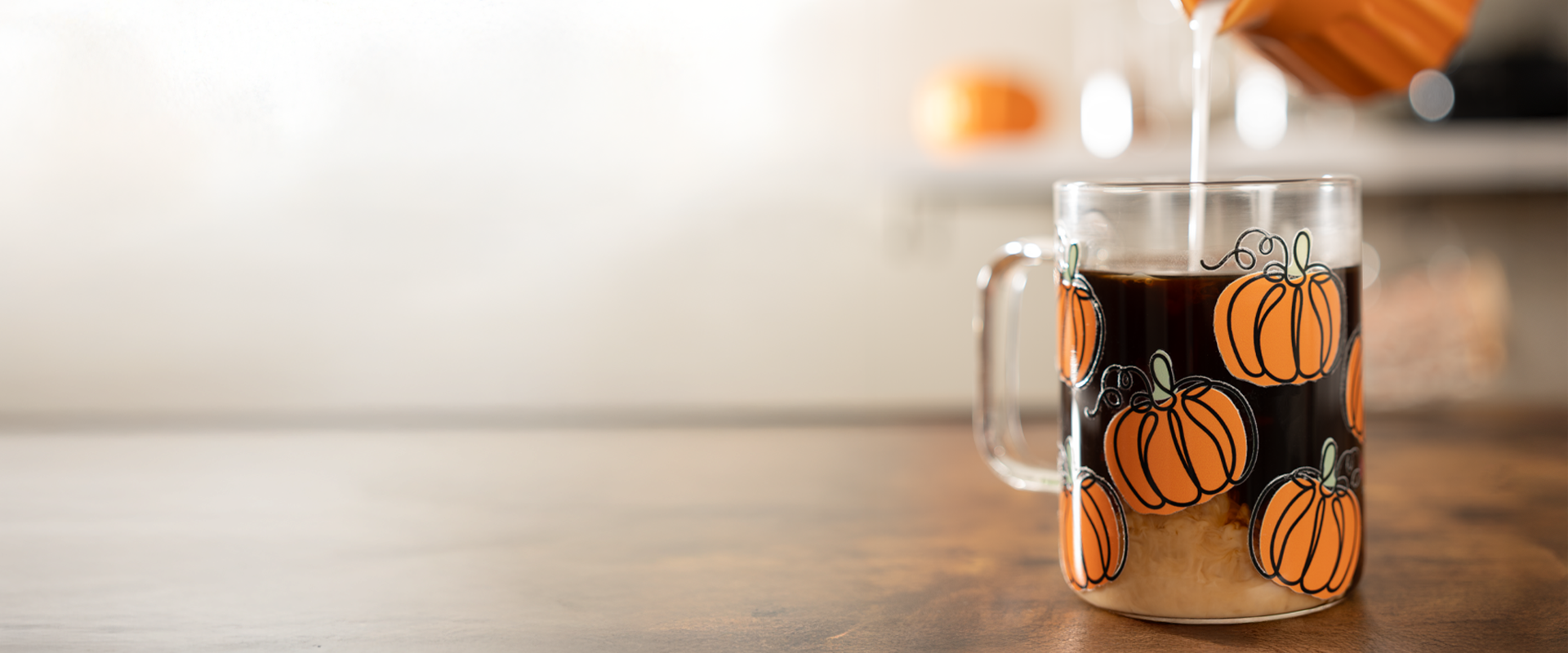A clear glass mug with a handle, decorated with a pattern of orange pumpkins, filled with a dark beverage and set on a wooden table. A wooden pitcher is pouring a light-colored liquid into the mug, with a softly blurred kitchen background.